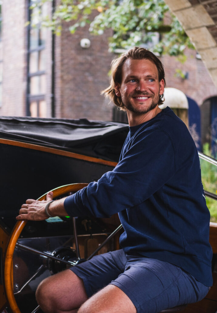 Smiling man in a navy sweater steering a small wooden boat outdoors.