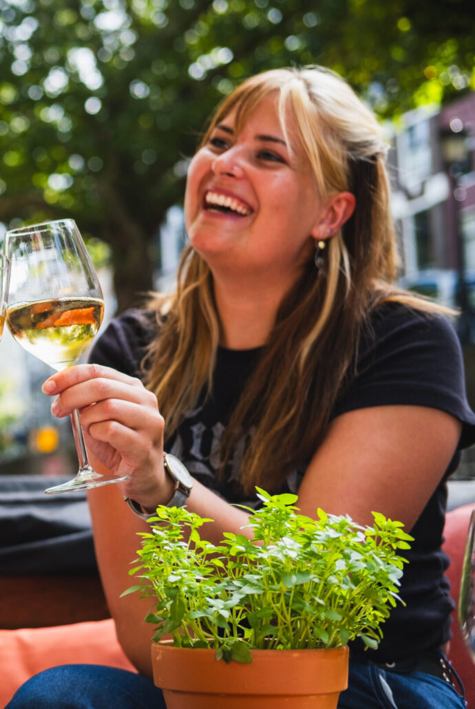 A smiling woman outdoors toasting with a glass of white wine, with a potted herb plant in the foreground and blurred greenery behind her.