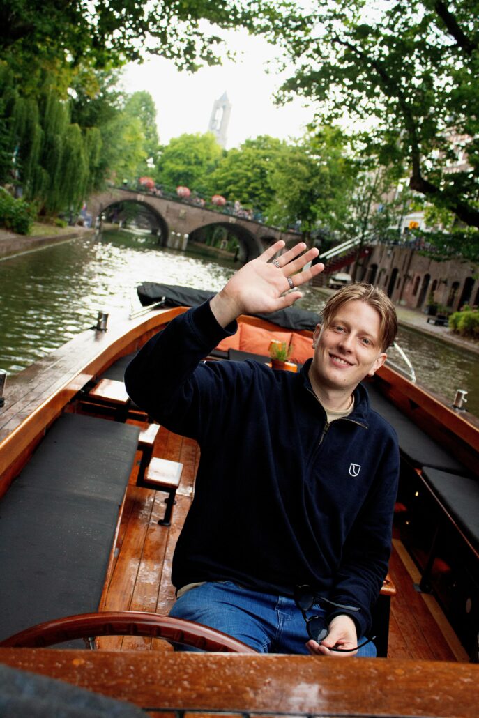 Smiling man on a wooden boat along a canal, waving at the camera with a brick arch bridge and green trees in the background.